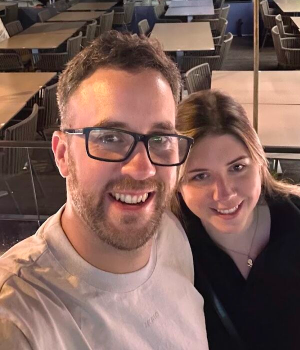 Smiling man with glasses and woman taking a selfie in a restaurant with tables and chairs in the background.