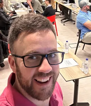 Smiling man with glasses and beard wearing a pink shirt in a busy indoor setting with people seated at tables in the background.