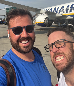 Two men smiling and posing for a selfie on an airport tarmac with a Ryanair airplane in the background.
