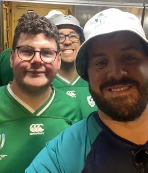 Three men smiling, wearing green sports jerseys and white hats in an indoor setting.