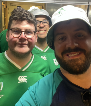 Three men smiling, wearing green sports jerseys and white hats in an indoor setting.