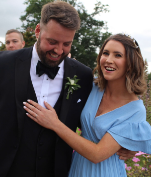 Smiling couple outdoors, woman in light blue dress with hand on man's chest, man in black tuxedo with bow tie and boutonniere.
