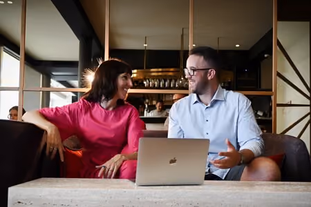 A man and woman sitting on a couch, smiling and conversing with a laptop on the table in front of them.