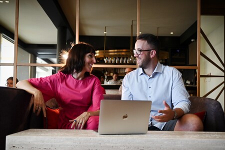 A man and woman sitting on a couch, smiling and conversing with a laptop on the table in front of them.