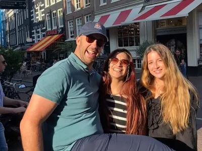 Three smiling people with sunglasses seated outdoors in a sunny urban area with cafes in the background.