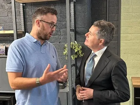 Two men engaged in a conversation indoors, one in a blue polo shirt and glasses, the other in a black suit with a blue tie.