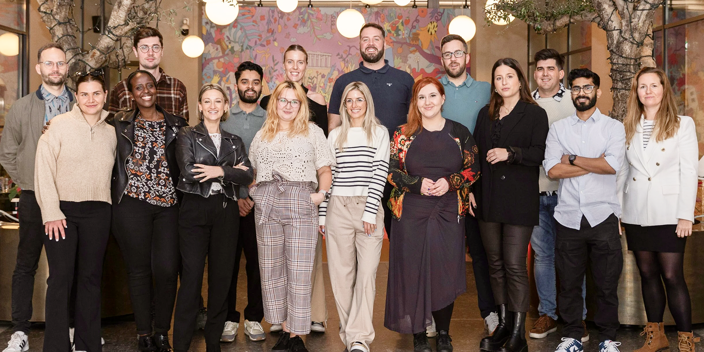 Diverse group of 16 people standing indoors in two rows, smiling, with trees and hanging globe lights in the background.