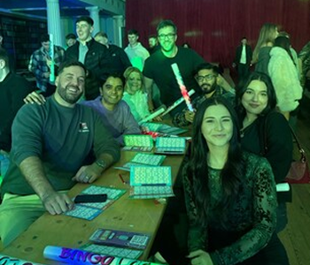 Group of seven adults smiling and posing at a table with wrapped gifts and bingo cards in a dimly lit indoor setting with red curtains.