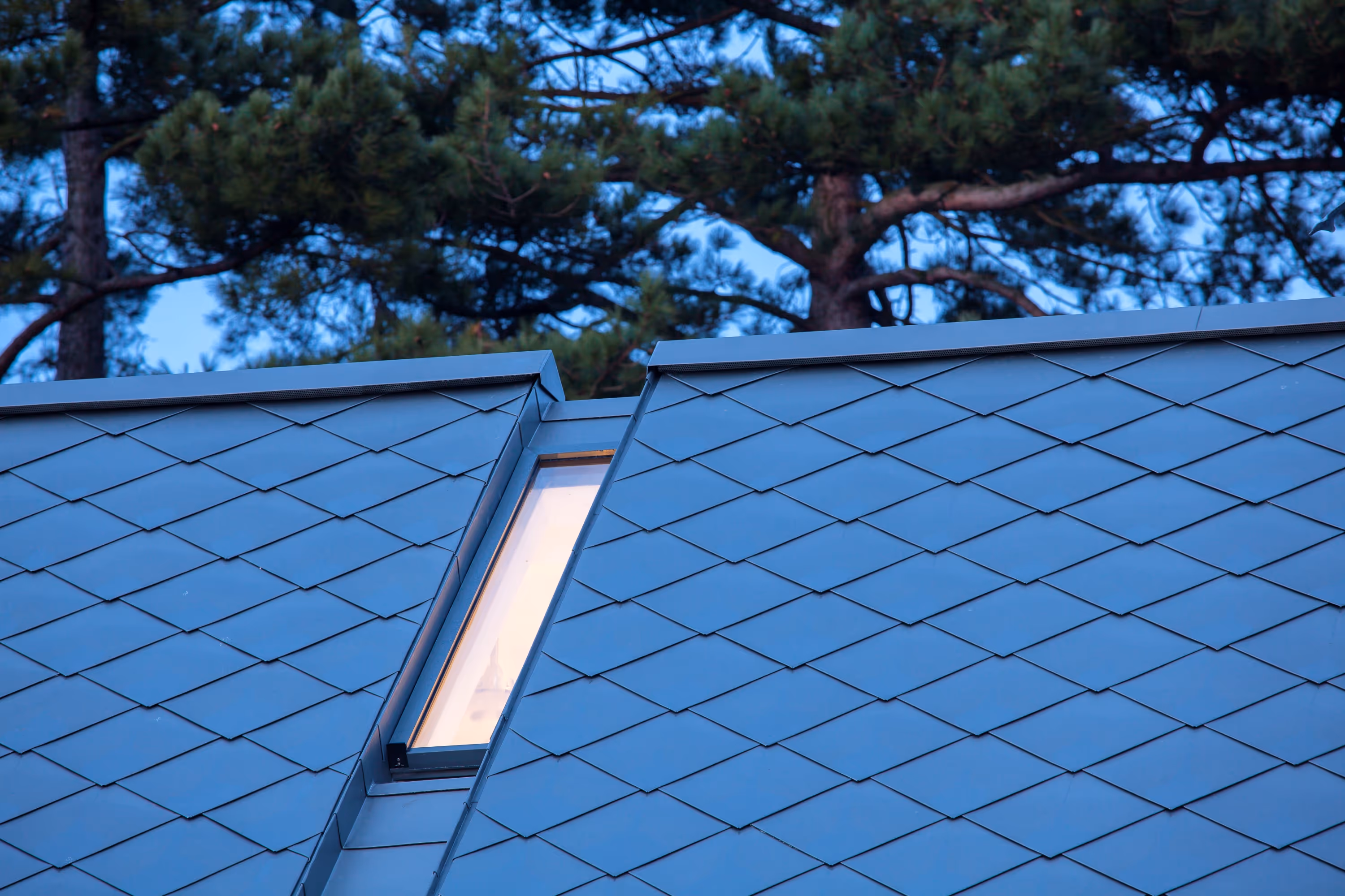 Close-up of a modern blue-tiled sloped roof with a narrow vertical window and trees in the background.