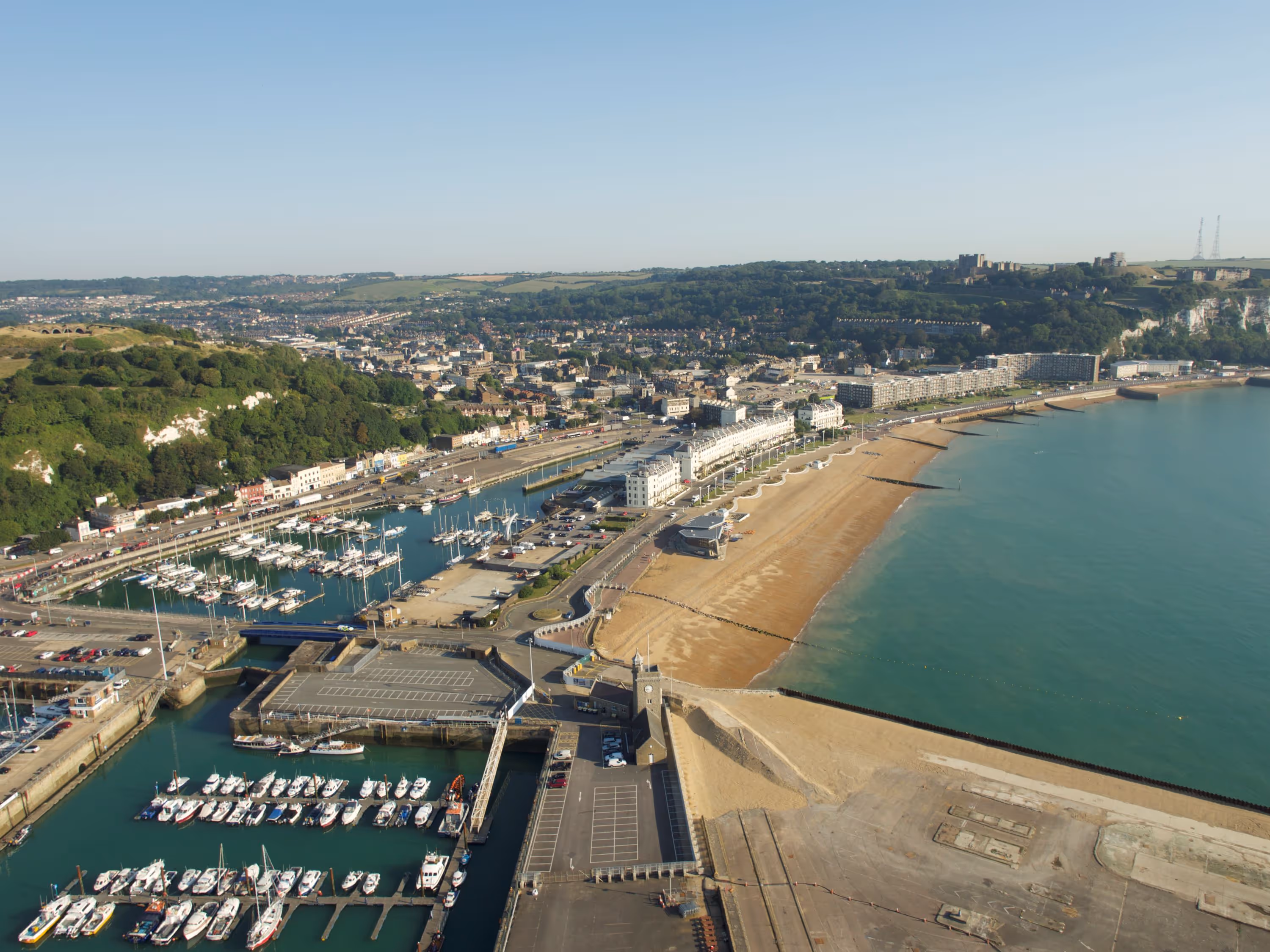 Aerial view of a coastal town with a marina full of boats, a sandy beach, and green hills in the background.