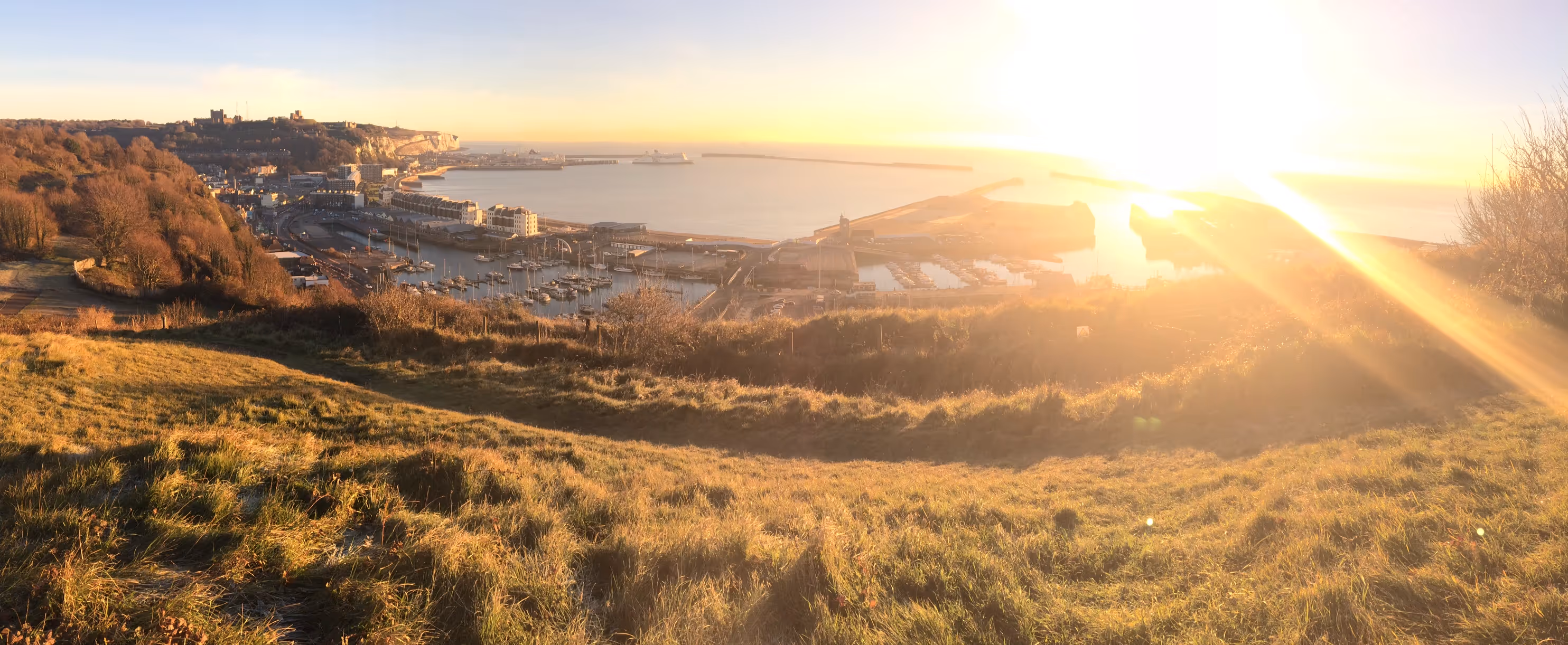 Sunset over a coastal town with a harbor filled with boats, grassy hillside in foreground, and a castle on a hill to the left.