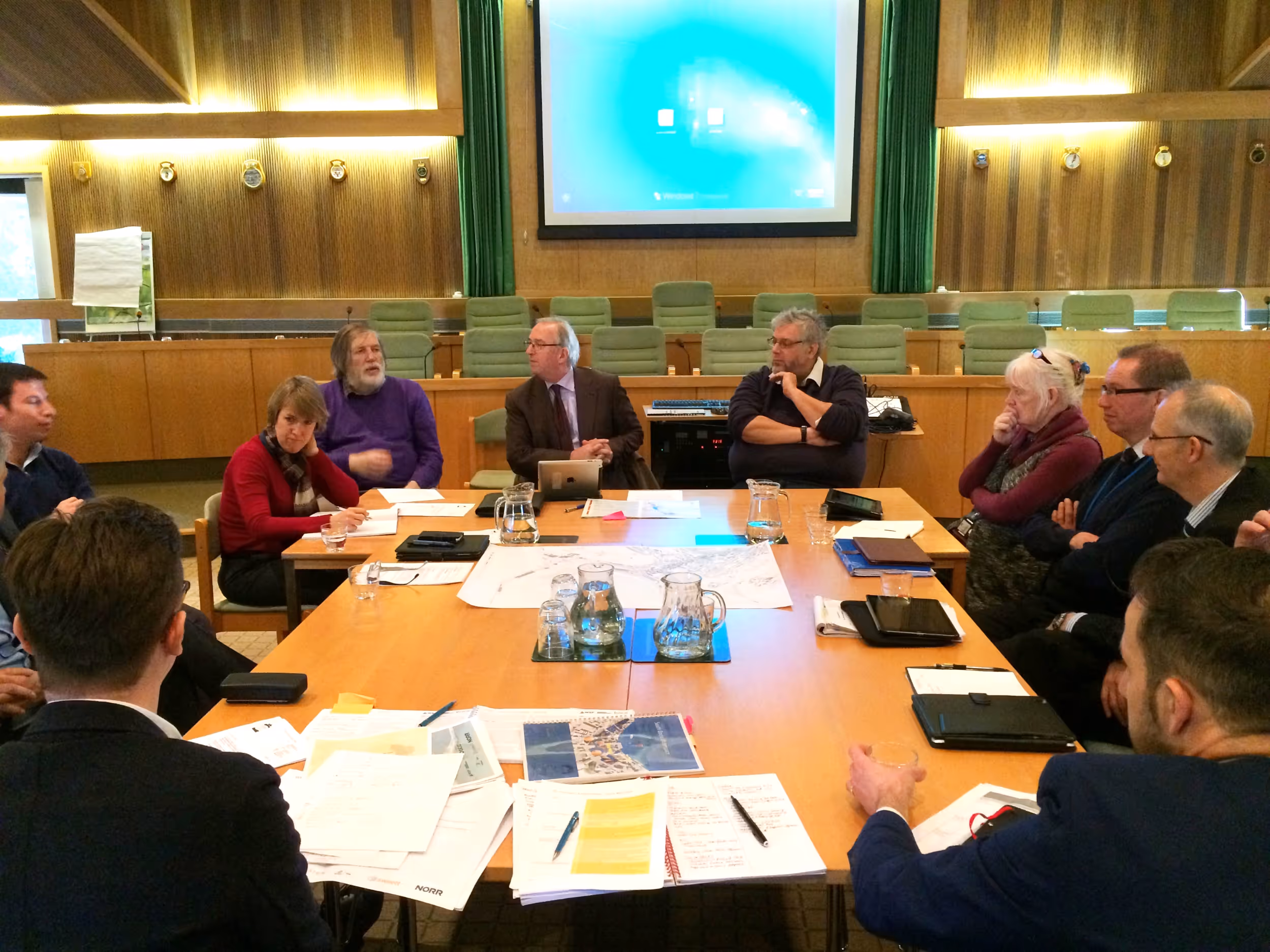 A group of people sitting around a conference table in a meeting room with documents, tablets, and water pitchers on the table.