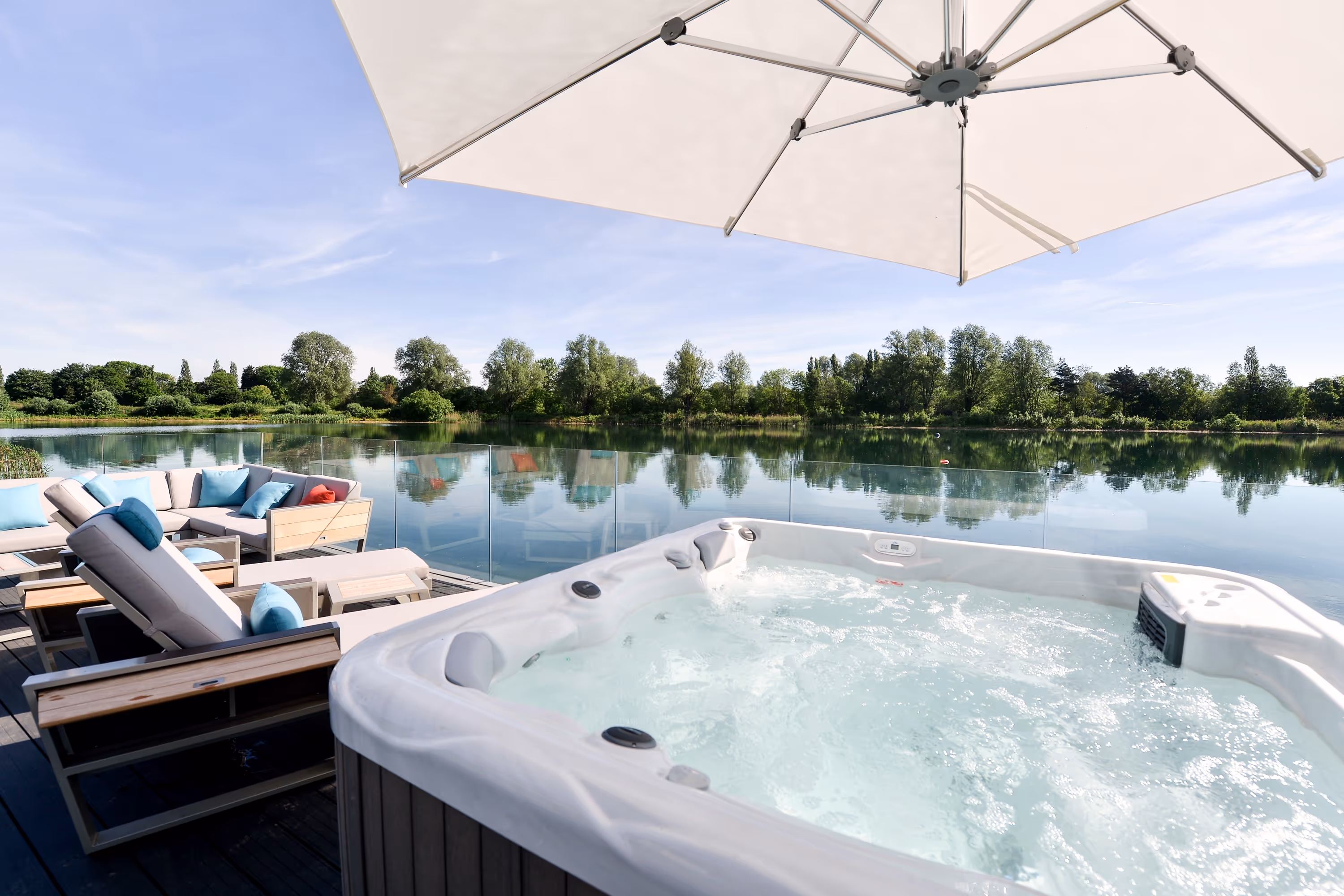 Outdoor hot tub with bubbling water beside lounge chairs and a large umbrella overlooking a calm lake and green trees.