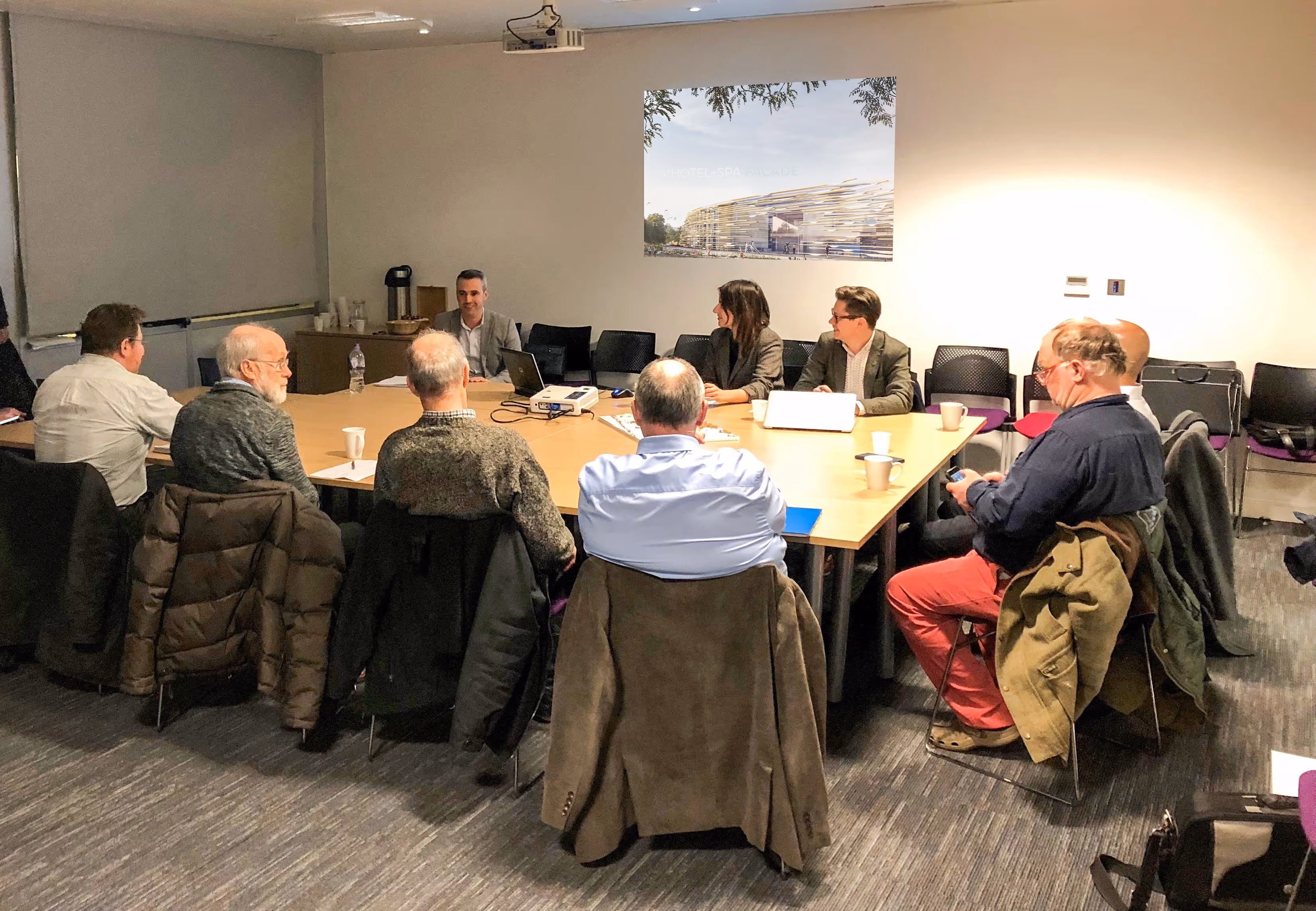 Meeting room with nine people seated around a rectangular table, some with laptops and coffee mugs.