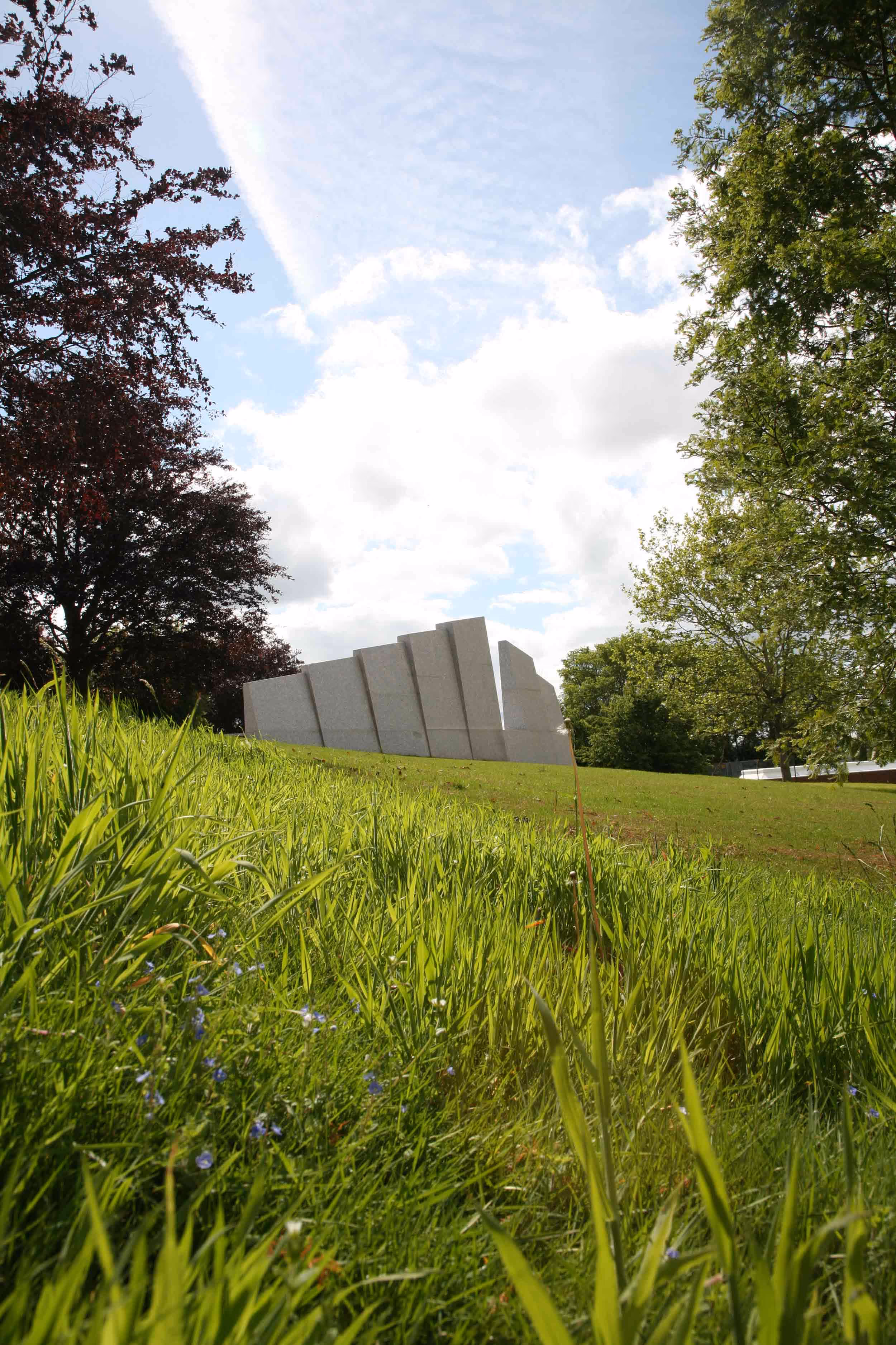 Green grassy hill with scattered trees and a modern angular stone monument under a blue sky with clouds.