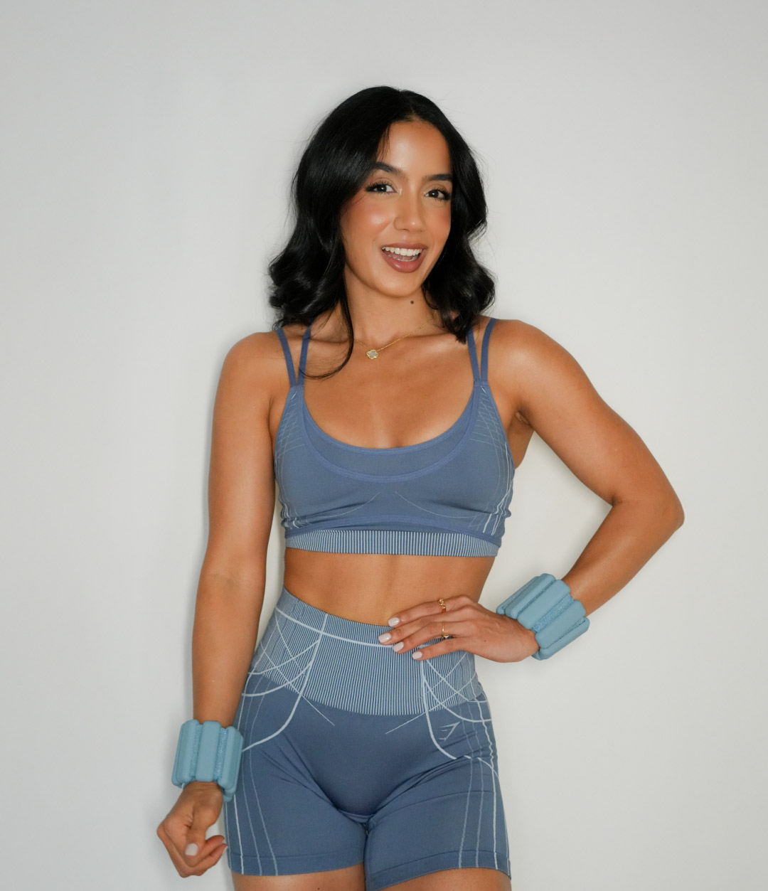 Mujer sonriente con cabello negro rizado, vestida con ropa deportiva azul y pesas en las muñecas, posando frente a un fondo blanco.