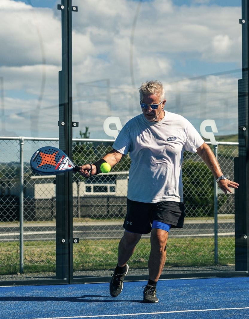Older man playing paddle tennis outdoors, hitting a ball with a paddle on a blue court.