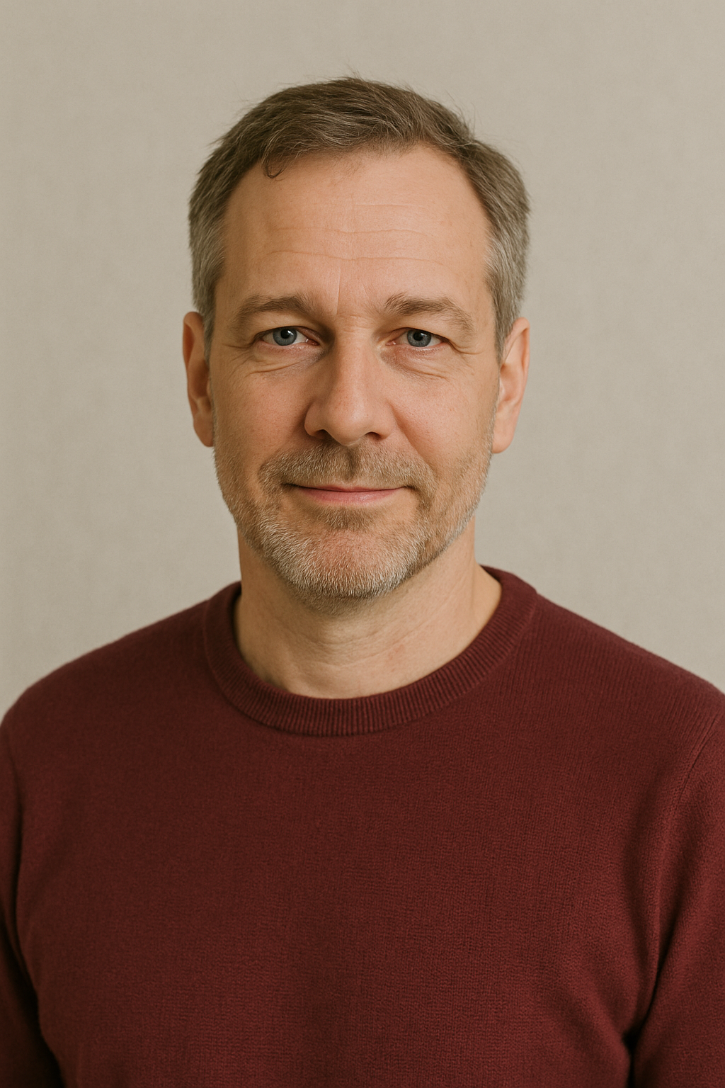 Middle-aged man with short graying hair and beard wearing a maroon sweater, smiling slightly against a beige background.
