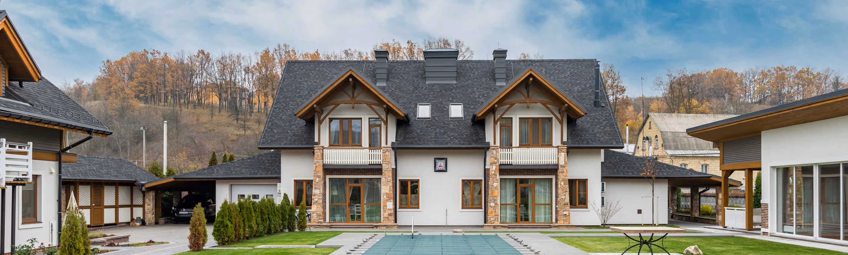 Сучасний двоповерховий будинок з мансардою, кам’яними оздобленнями та темним дахом на фоні осіннього пейзажу.