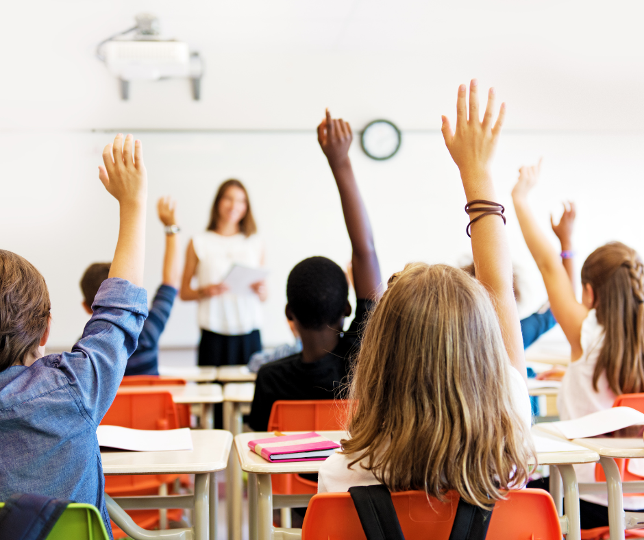 Classroom with several students with their hands raised and a teacher standing at the front of the classroom.