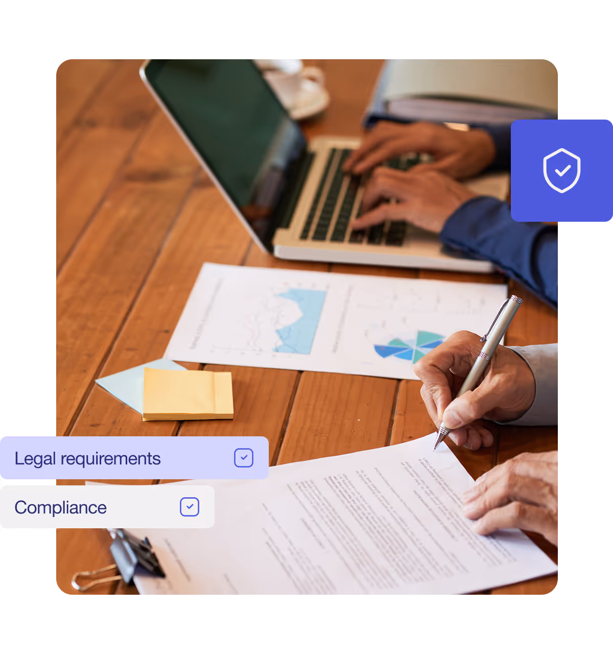 Person signing a document on a wooden desk with another person typing on a laptop, alongside business charts and sticky notes.