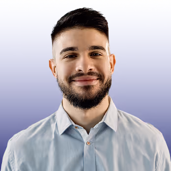 Smiling young man with beard and short dark hair wearing a light blue button-up shirt against a purple background.