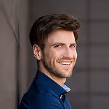 Smiling man with short brown hair and stubble wearing a dark blue shirt against a neutral background.