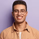 Smiling young man with glasses and short curly hair wearing a tan button-up shirt against a lavender background.