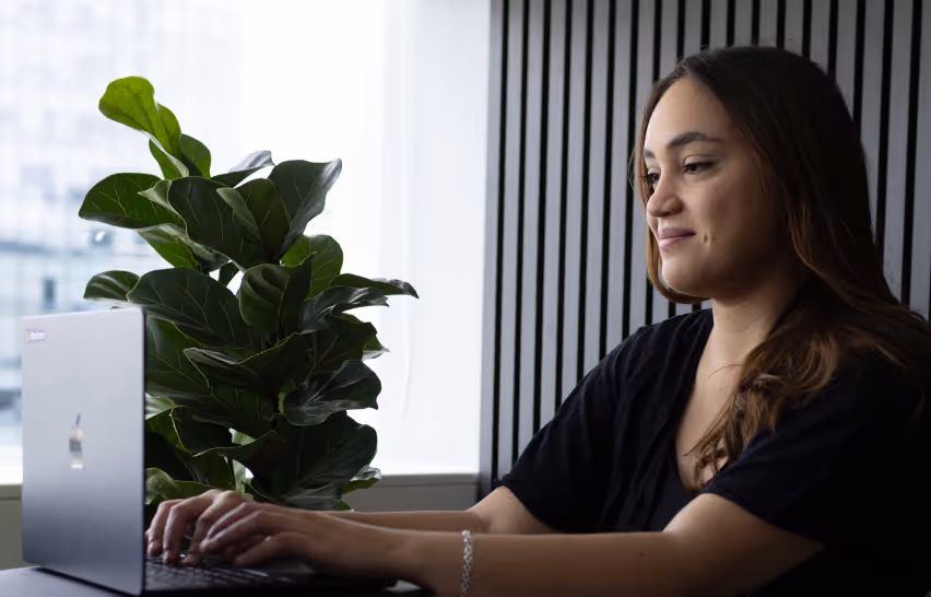 Woman with long hair typing on a laptop near a large green leafy plant by a window.
