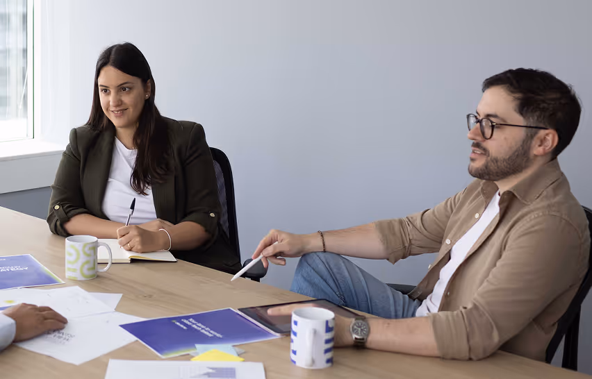 Two colleagues sitting at a table during a meeting, one holding a pen and notebook, the other gesturing with a pen.