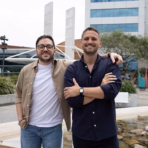 Two smiling men standing outdoors by a water feature in front of a modern building, one with glasses and beige shirt, the other with arms crossed wearing a dark blue shirt.