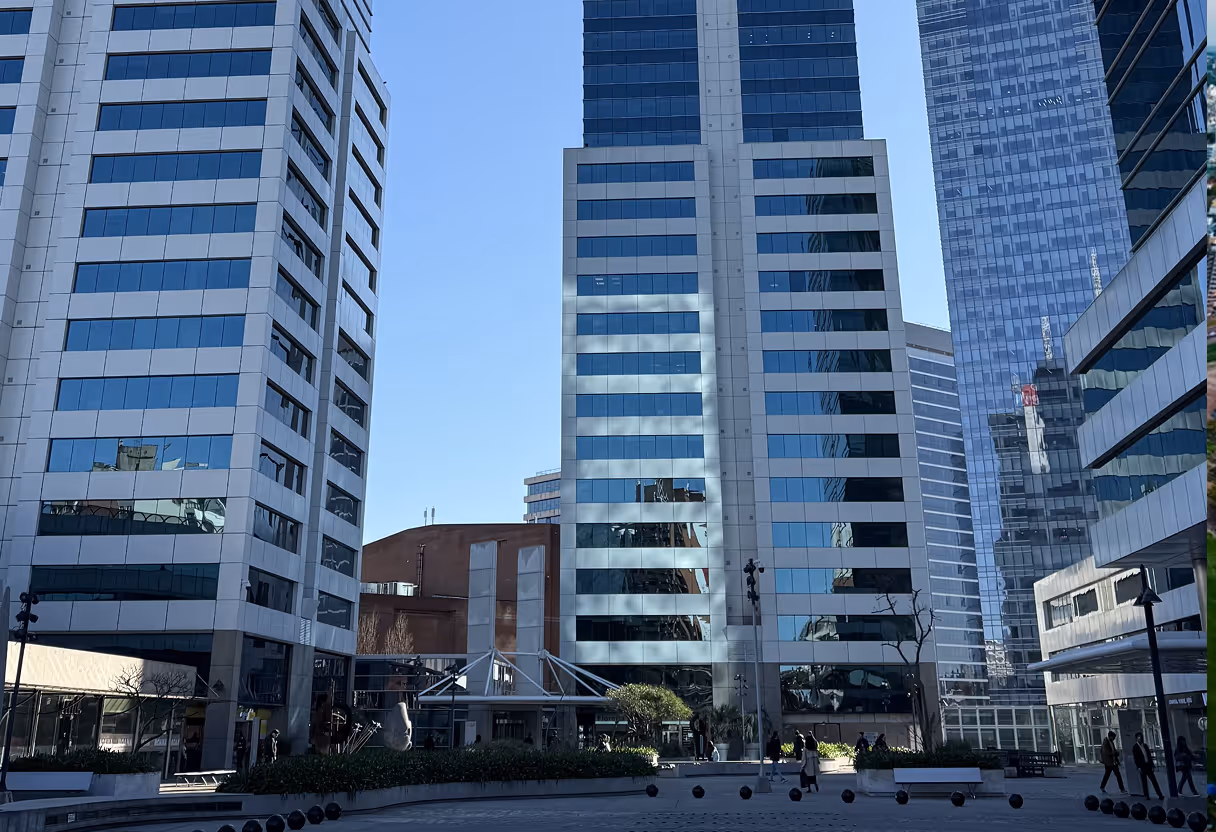 Modern office buildings surrounding a plaza with benches and small trees under a clear blue sky.