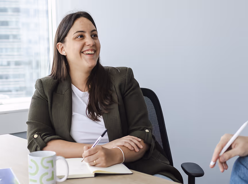 Smiling woman in blazer seated at a desk, holding a pen over an open notebook during a meeting.