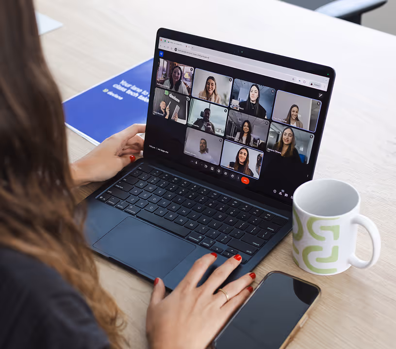 Person using a laptop on a desk with a video conference showing nine participants, next to a mug and a smartphone.