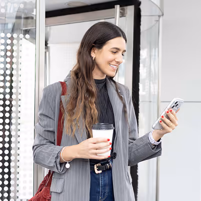 Woman in a gray pinstripe blazer holding a coffee cup and smiling at her phone near a glass door.
