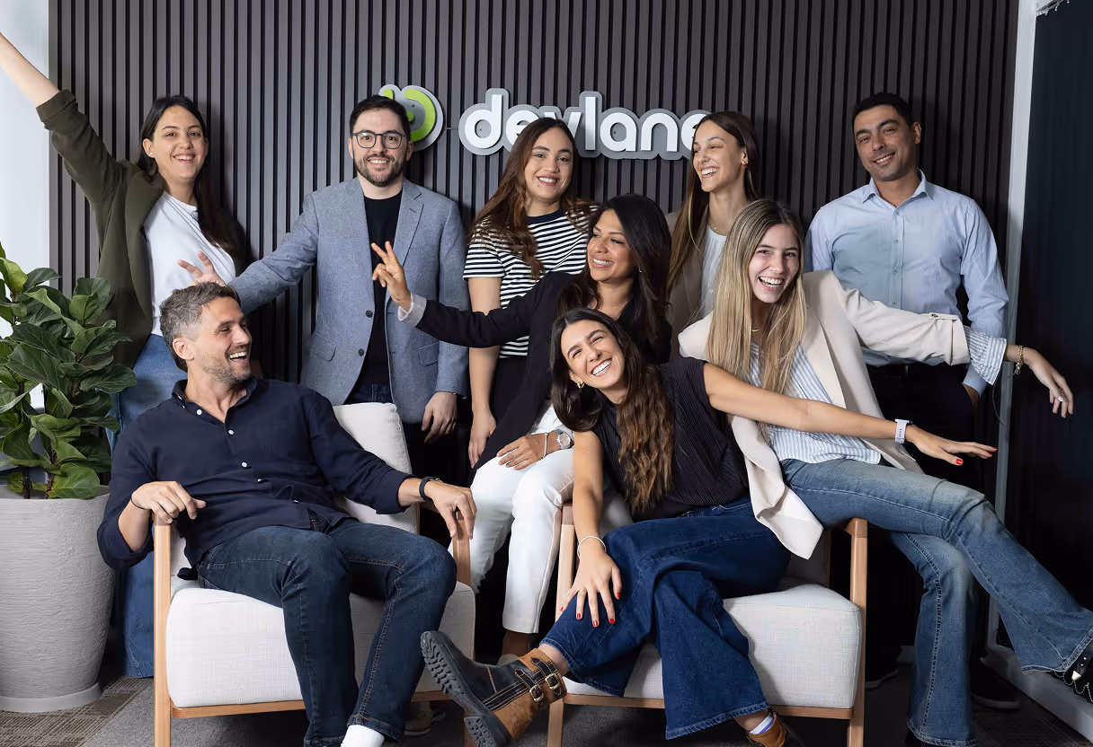 Group of nine young professionals smiling and posing cheerfully in an office setting with a potted plant and 'devland' logo on the wall behind them.