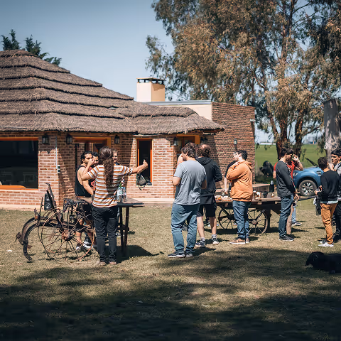 Group of people socializing outdoors near rustic brick buildings with thatched roofs on a sunny day.