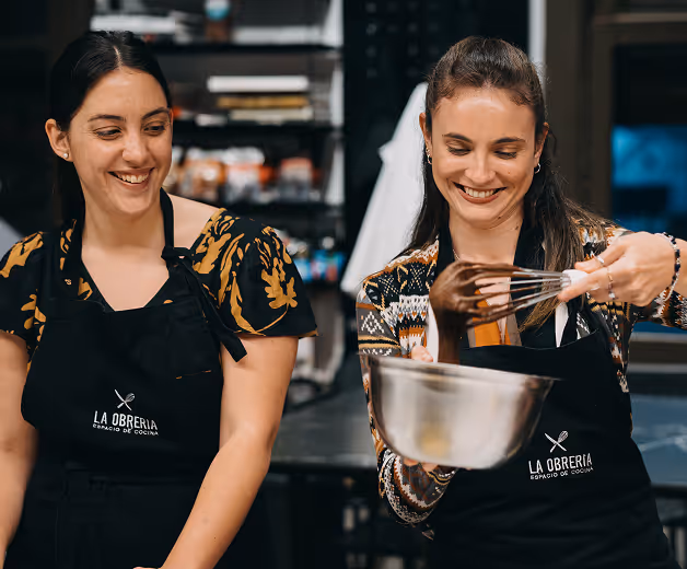 Two women wearing La Obreria aprons, one stirring chocolate in a mixing bowl with a whisk while the other smiles watching.