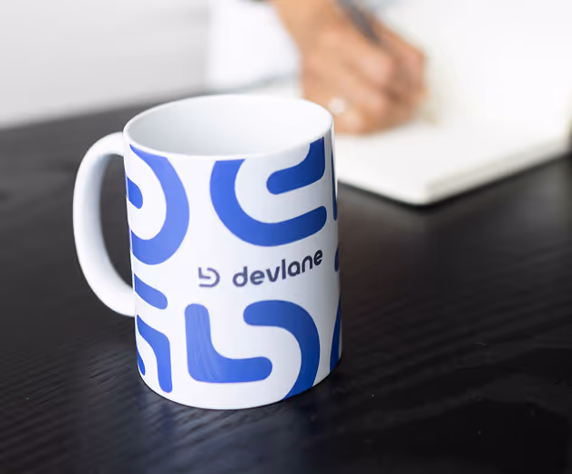 White ceramic mug with blue abstract patterns and the word 'devlane' on a black wooden table, with a blurred hand writing in a notebook in the background.