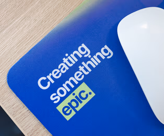 Blue mouse pad on a wooden desk with text 'Creating something epic.' and a white computer mouse on it.