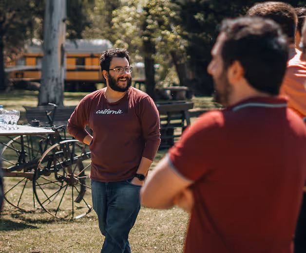 A man in a maroon California sweatshirt and glasses smiling outdoors while talking to two other men.
