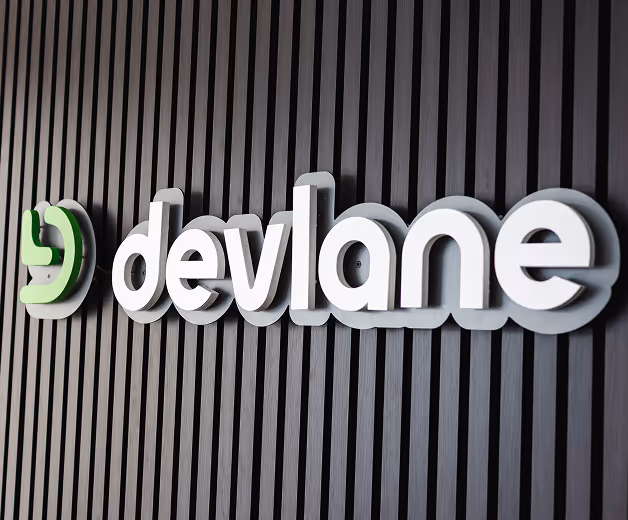 3D white and green Devlane logo mounted on vertical black slatted wall.