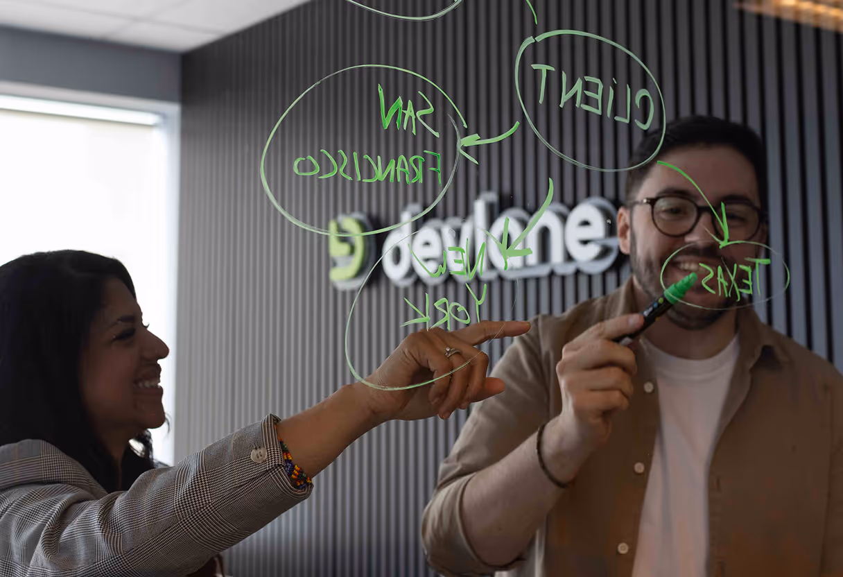 A man and woman collaborating, writing with a green marker on a glass board with business-related words and arrows.