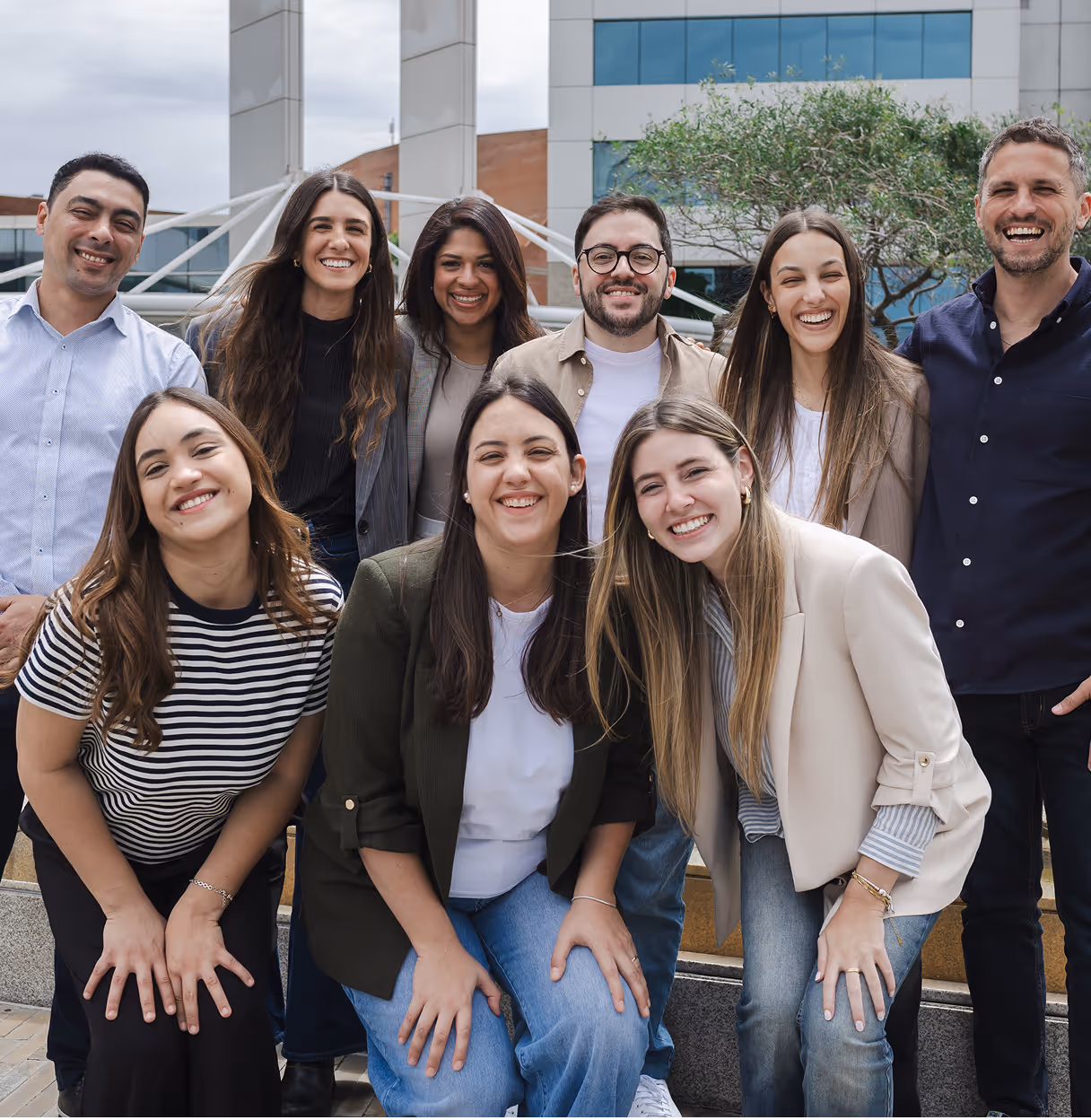 Group of eight diverse young adults smiling outdoors in business casual attire with a modern office building in the background.