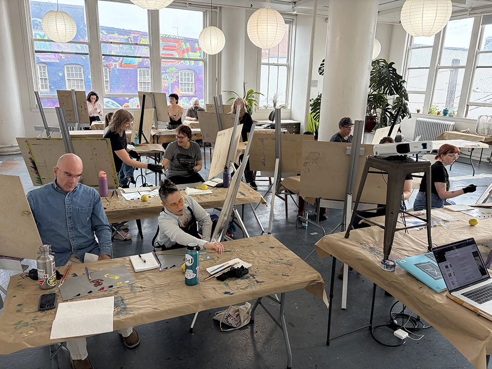 A painting class in a bright loft studio with tall windows and paper lanterns, students working at easels and long tables.