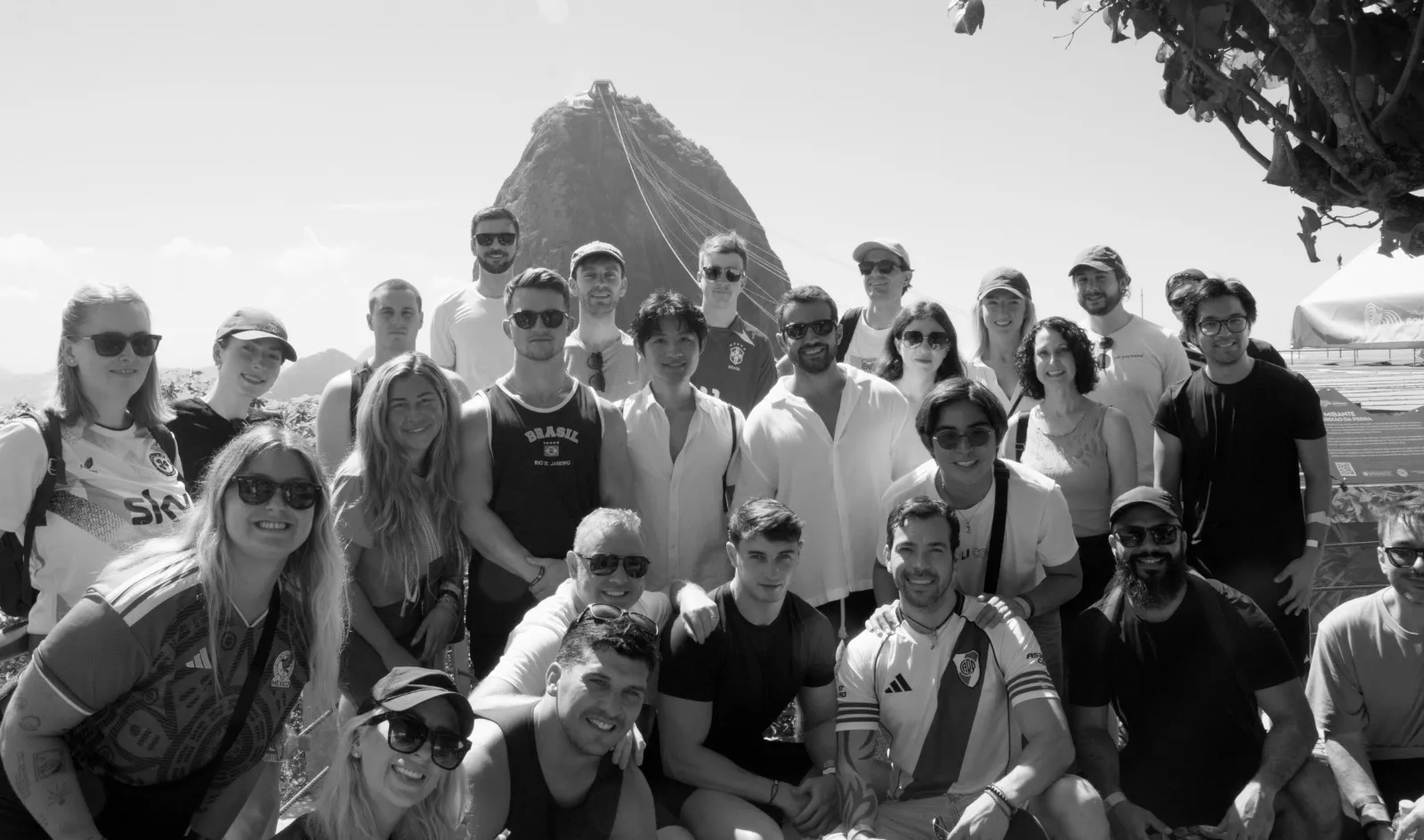 A large group of people posing outdoors with a mountain and cable cars in the background.