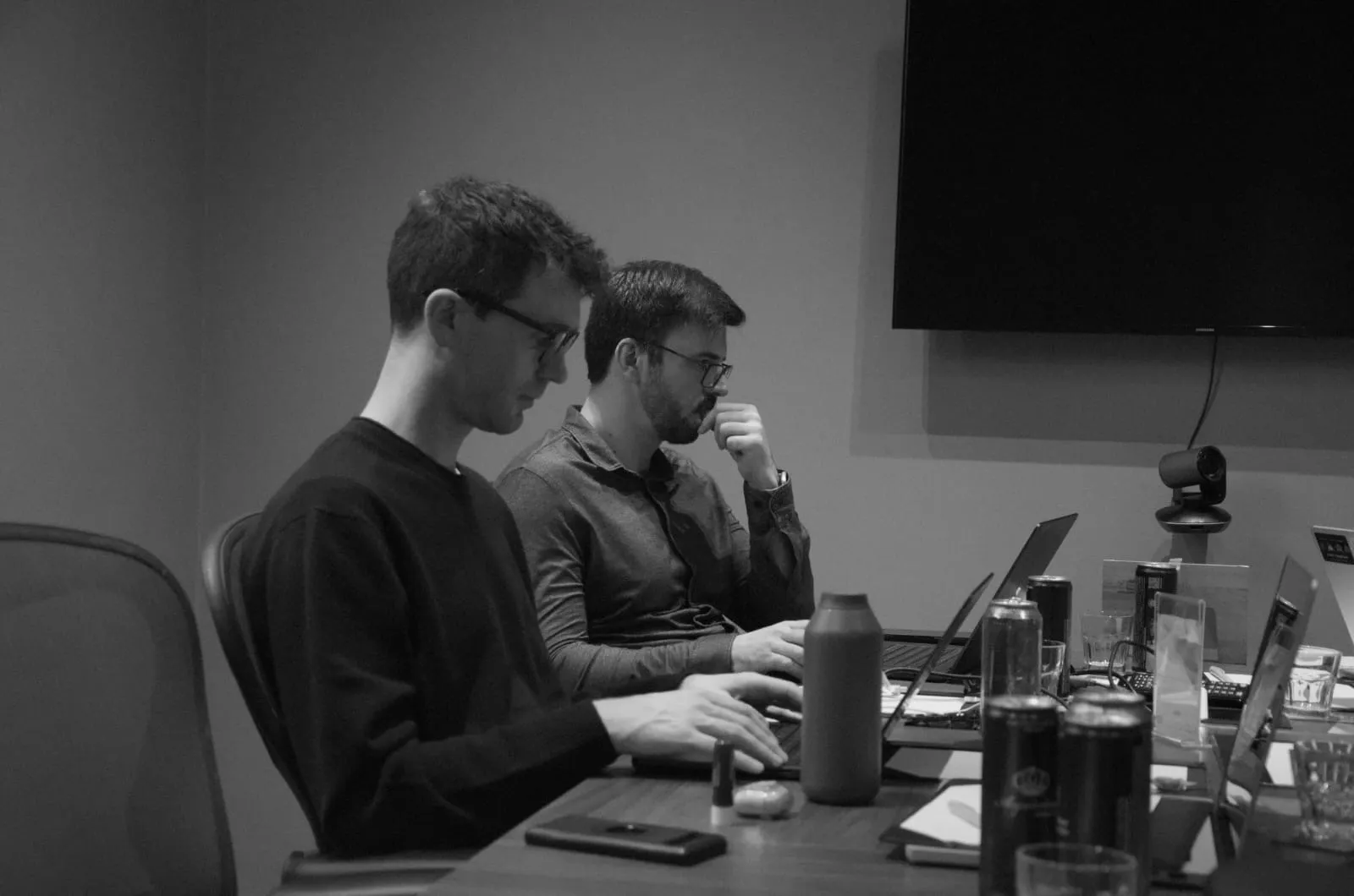 Two men wearing glasses working on laptops at a table with various drinks and notebooks.