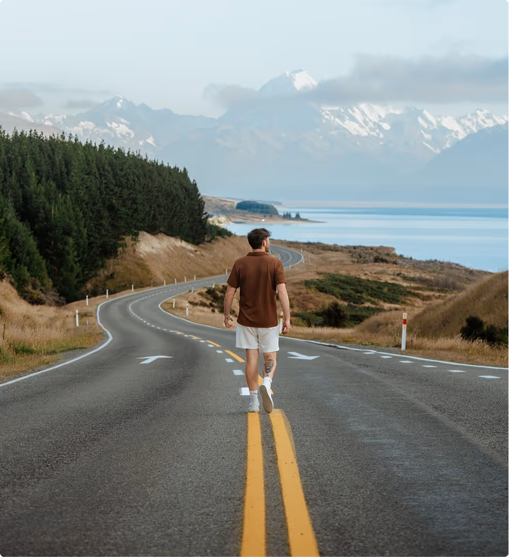 Man in brown shirt and white shorts walking on a winding road with trees, hills, lake, and snow-capped mountains in the background.