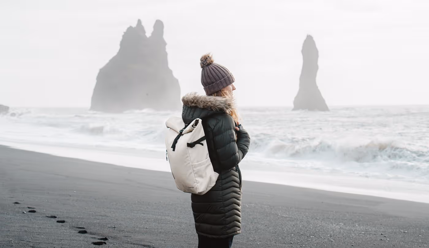 Person wearing a winter coat and beanie with a backpack standing on a black sand beach facing the ocean and rock formations.