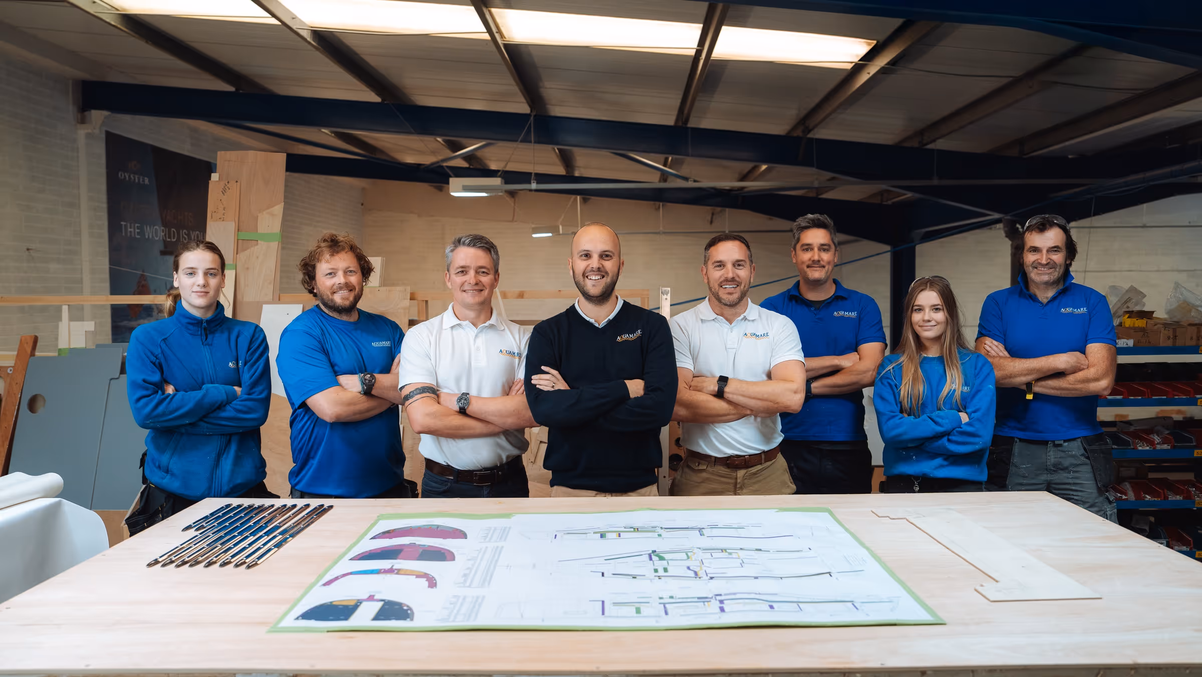 Eight team members standing behind a table with blueprints and tools in a workshop, wearing Aquamare branded clothing.